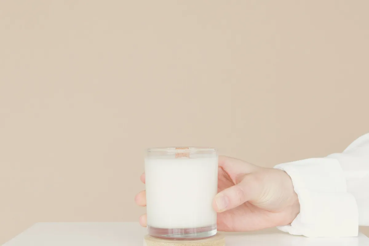 A hand resting around a small glass of milk on a pale table against a warm beige wall.