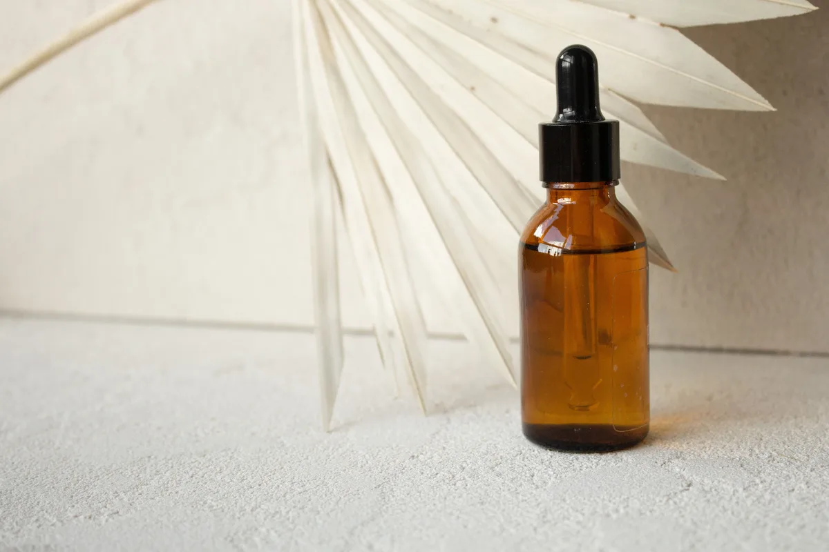 A small amber glass dropper bottle standing on a pale cream surface beside a dried palm leaf.