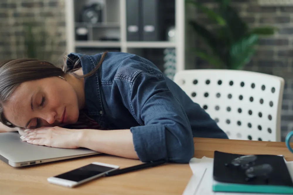 A woman asleep at her desk during the day, head resting on her arm beside a laptop.