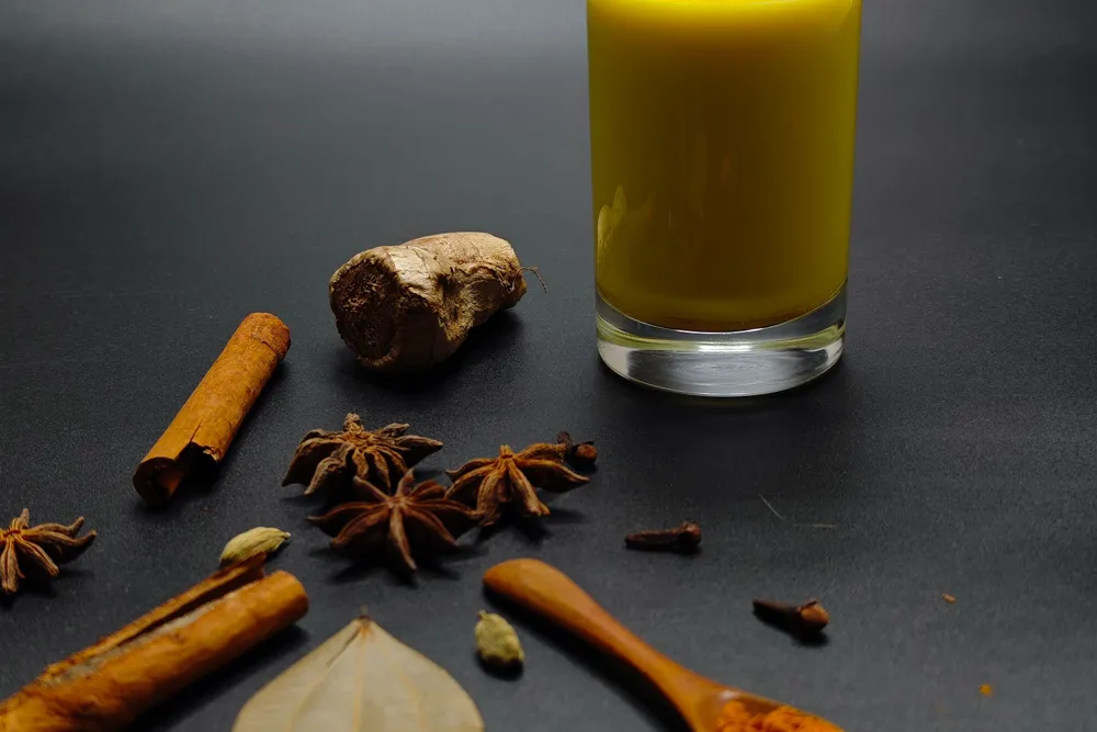 A glass of warm spiced golden milk surrounded by cinnamon, star anise, ginger and cardamom on a dark surface.