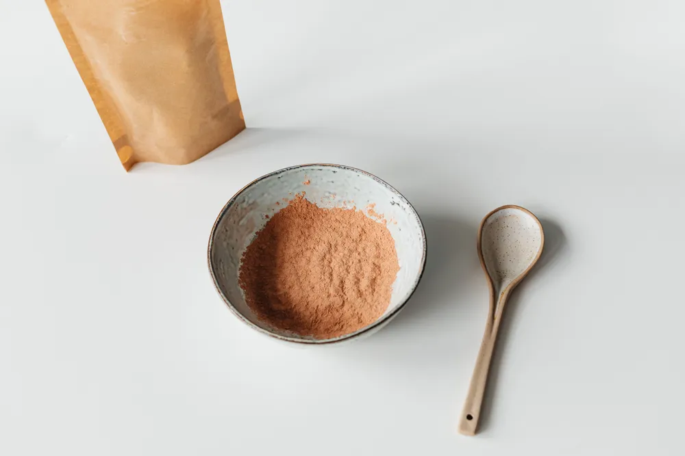 A small bowl of tan-brown ashwagandha root powder beside a wooden spoon and a kraft pouch.
