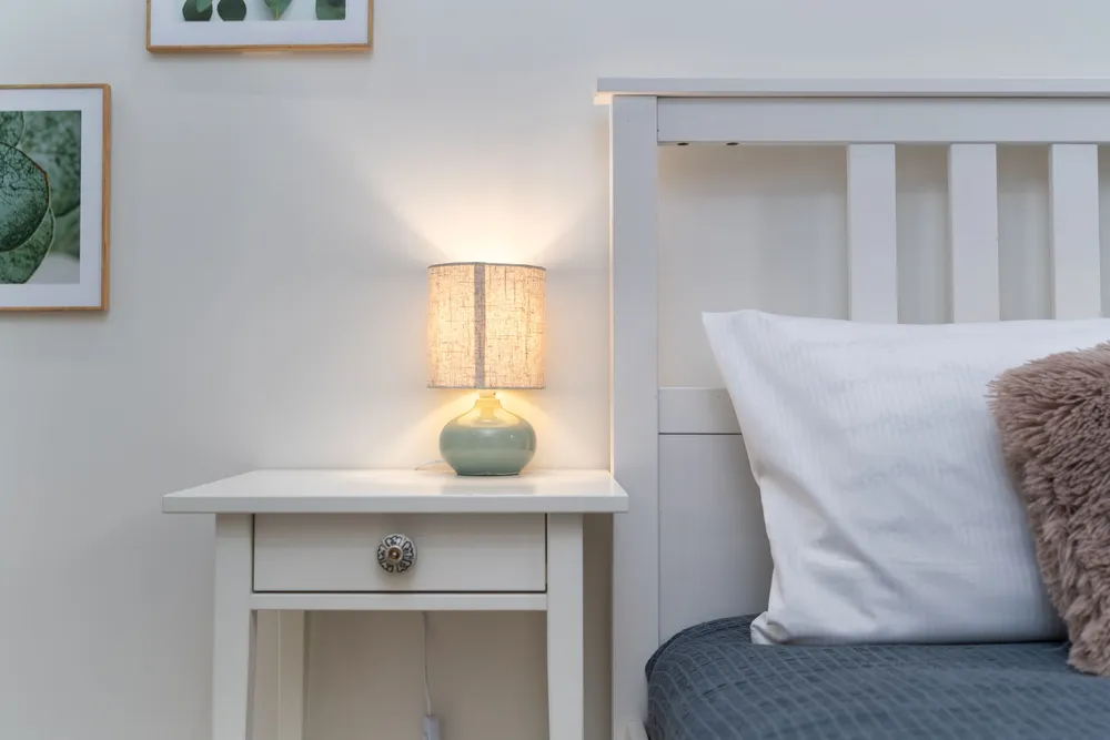 A small bedside table with a lit lamp beside a made bed in a quiet bedroom.