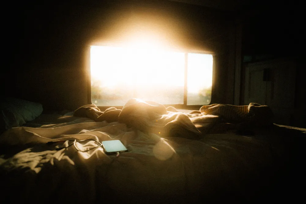 A rumpled bed with a smartphone on the sheets and bright light glowing behind it.
