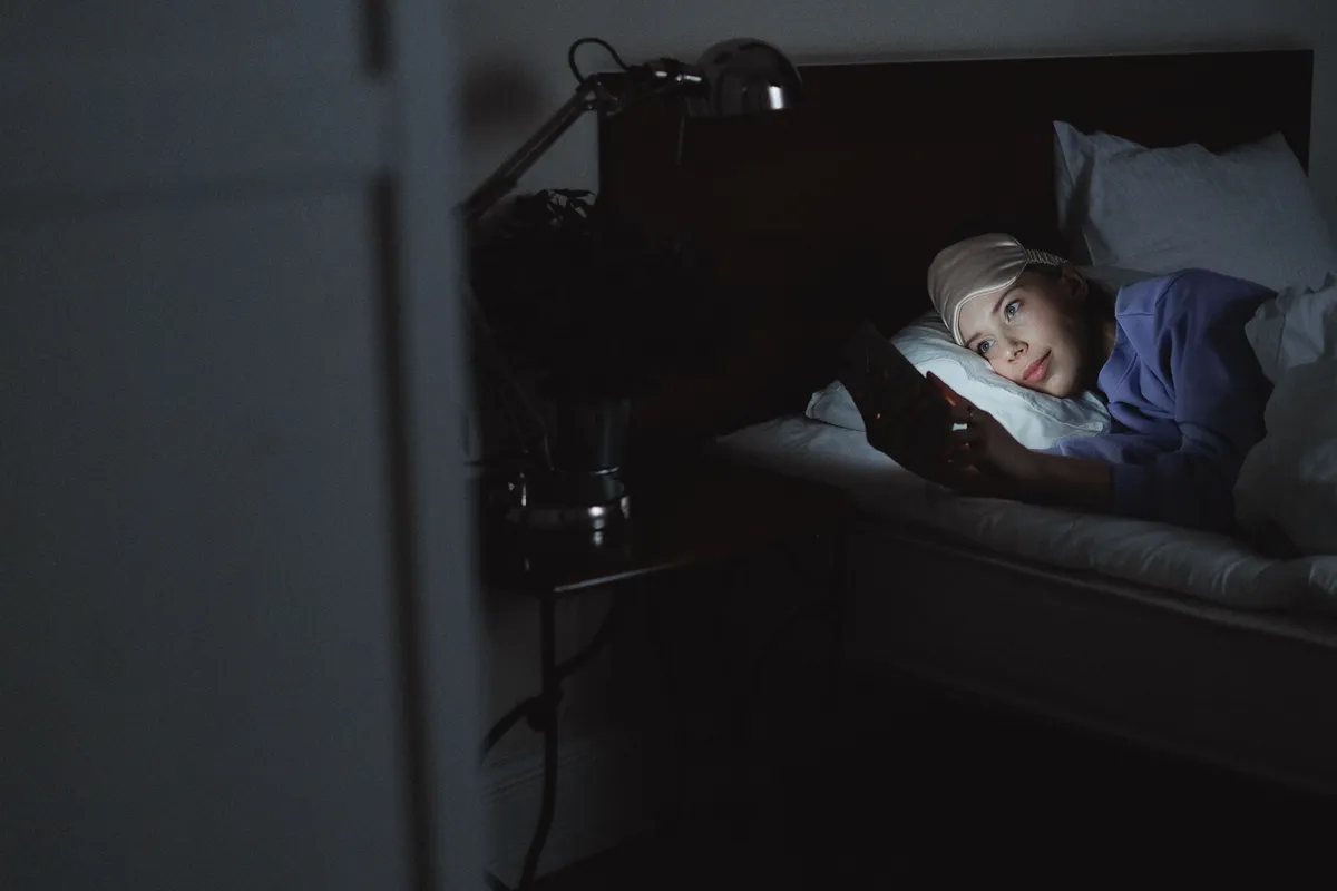 A woman in a dark bedroom, her face lit by the glow of a smartphone screen.