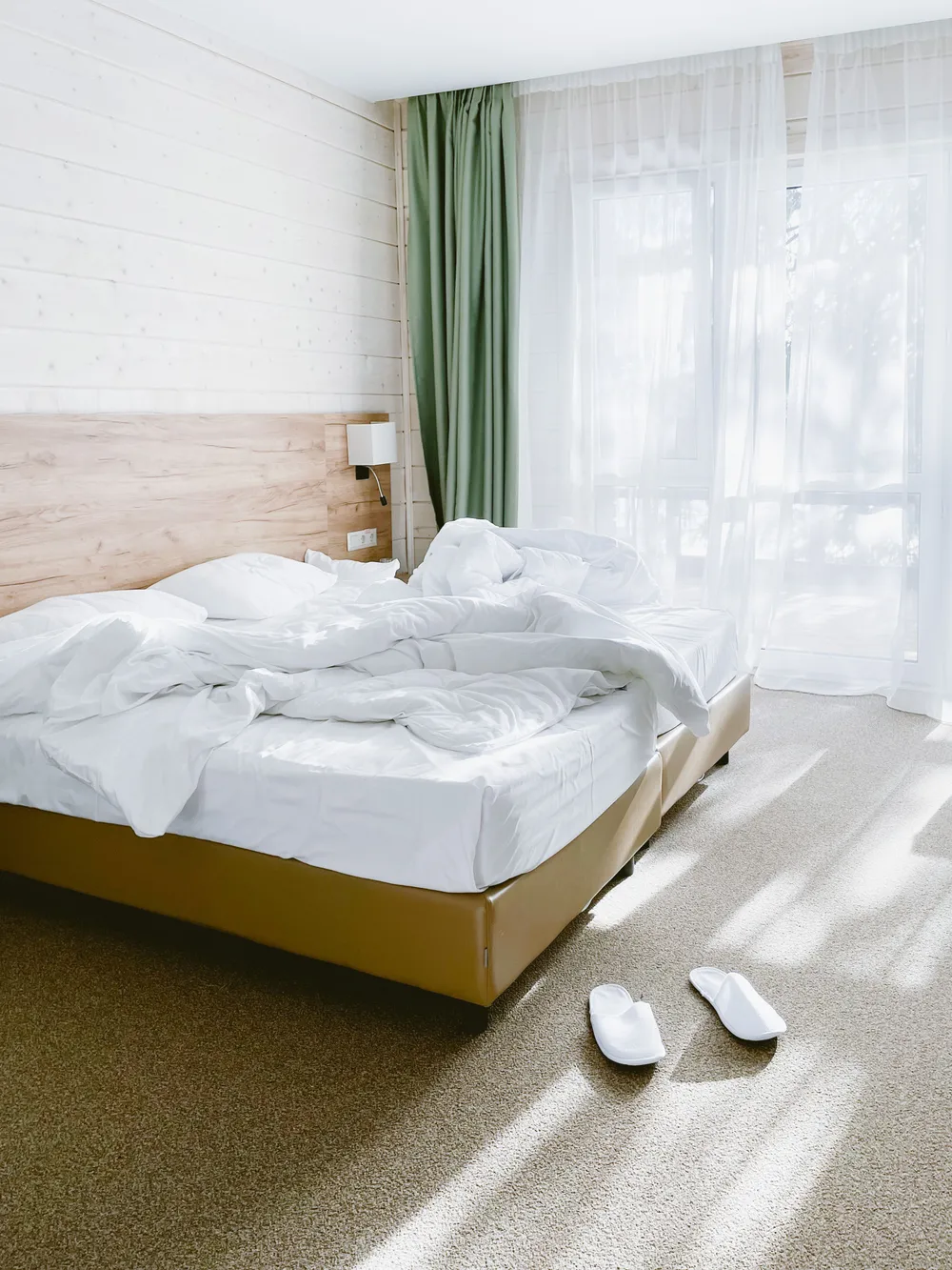 A bright, airy bedroom with light bedding, sheer curtains and a soft green drape by large windows.