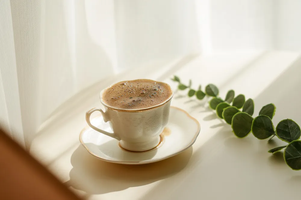 A cup of coffee on a saucer beside a window, in soft morning light with long shadows.