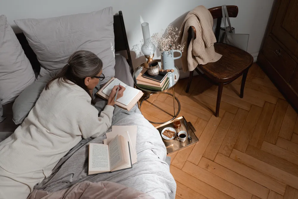 A mature woman in glasses writing in a notebook in bed by lamplight at night.