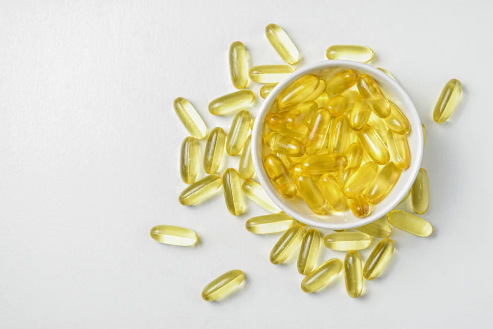 Golden fish oil capsules spilling from a small white bowl and arranged in a circle on a white surface.