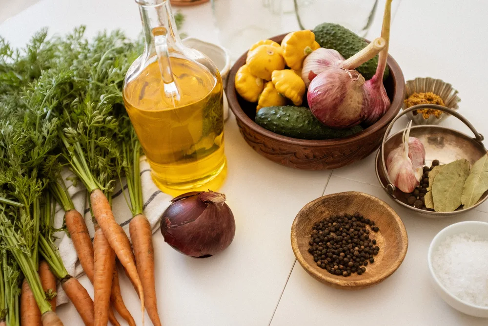 Fresh carrots, courgettes, garlic, peppercorns and a bottle of olive oil arranged on a pale kitchen table.