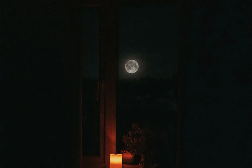 A full moon visible through a dark window, with a warm candle glowing on the sill inside the room.