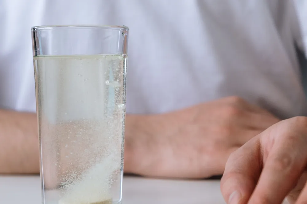 A glass of water on a pale table with a person's hands resting nearby.