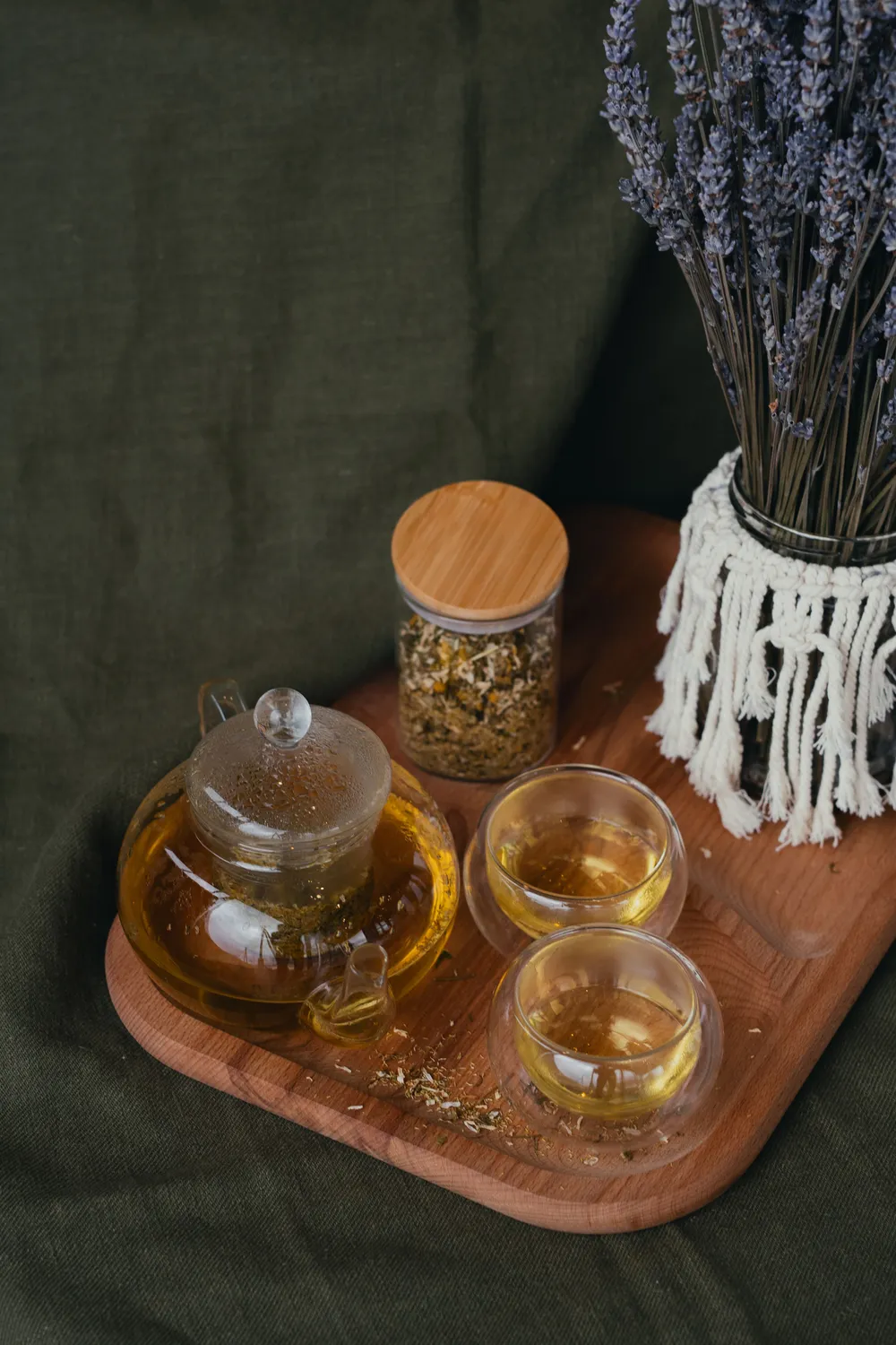 A glass teapot and cups of herbal tea beside a jar of dried chamomile and a bunch of dried lavender on a wooden tray.