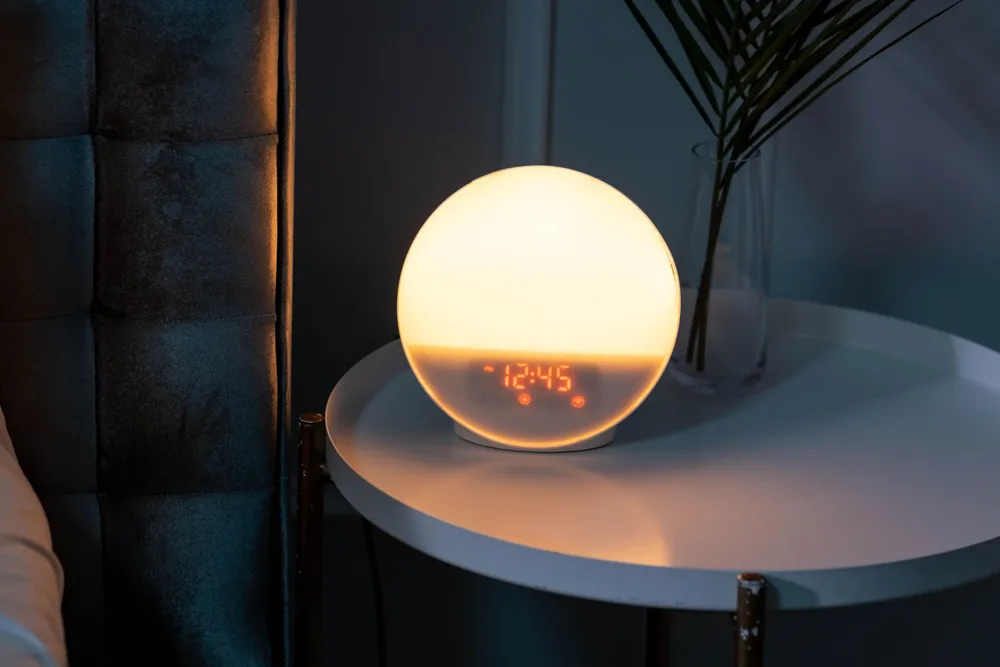 A round glowing bedside clock showing 12:45 on a small table beside a bed in a dark room.