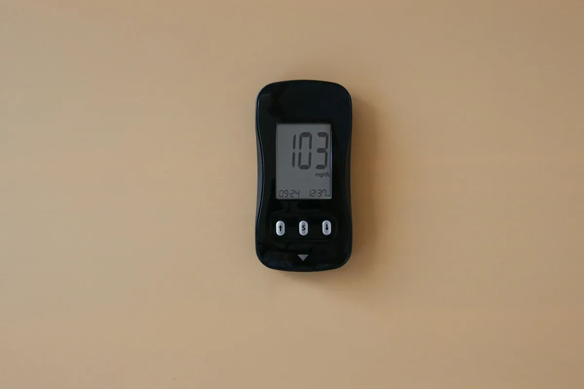 A small black blood glucose meter on a warm beige wall showing a reading of 103 mg/dL.