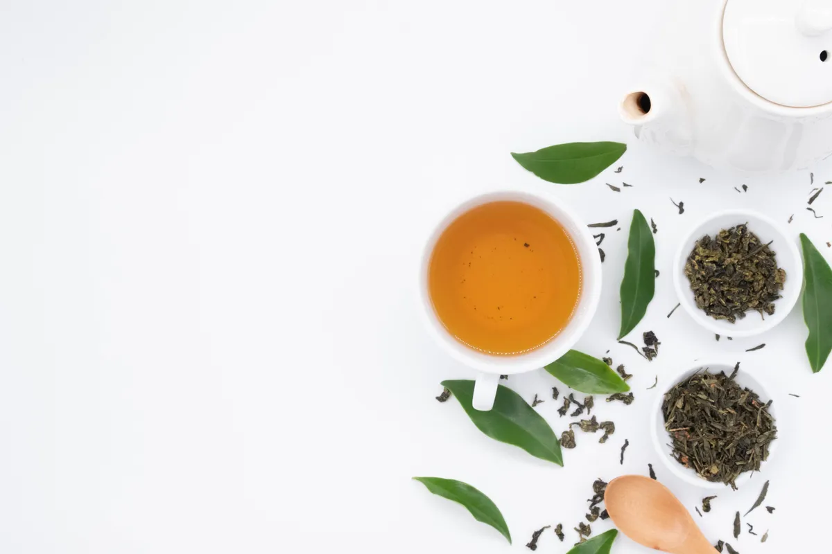 A cup of tea beside bowls of dried loose tea leaves and fresh green leaves on a white surface.