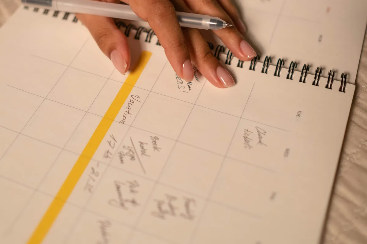 A hand holding a pen over an open paper planner, with a run of days marked in yellow highlighter.