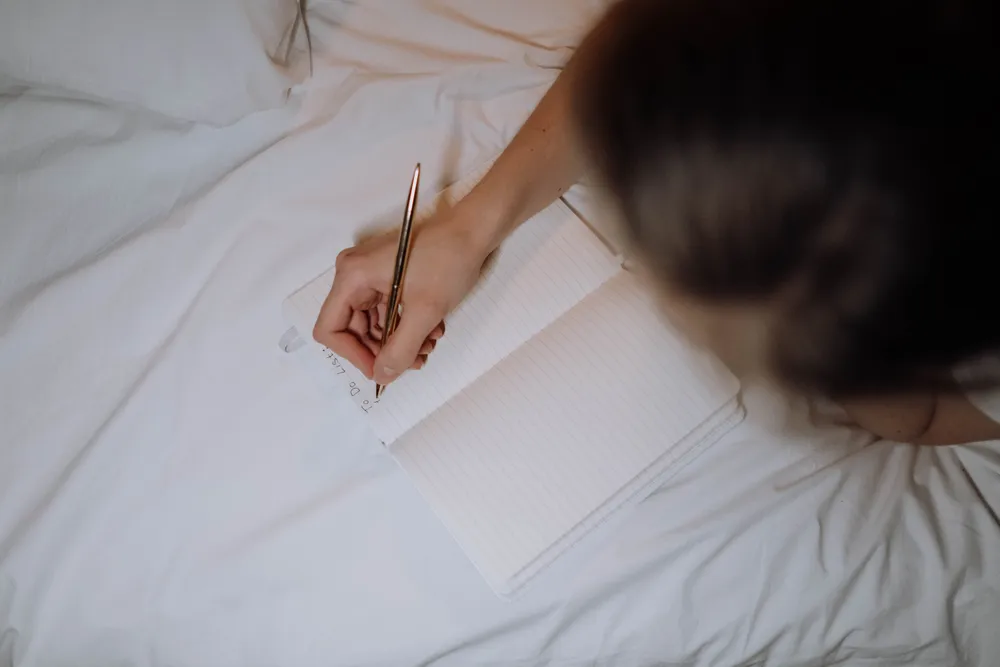 A hand writing notes in a lined notebook resting on white bed linen, seen from above.