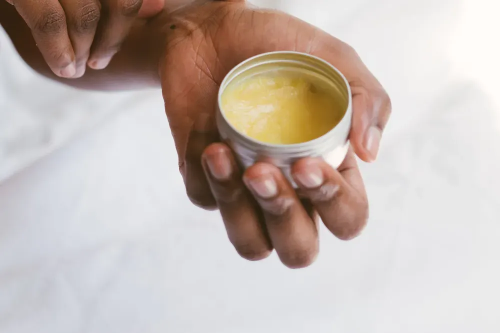 Hands holding an open metal tin of pale yellow cream against a white background.
