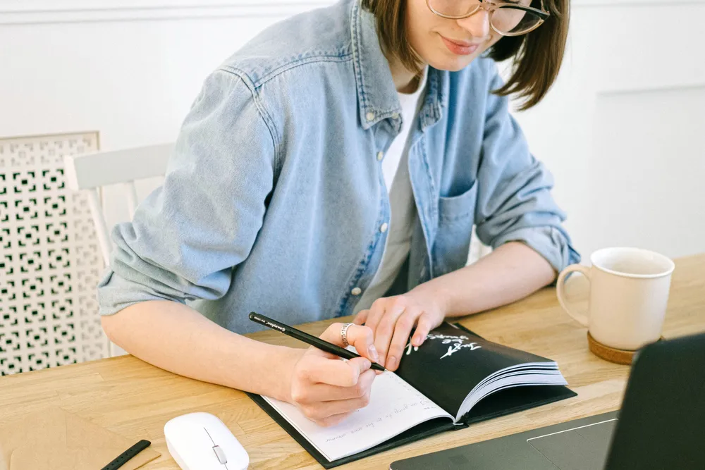 A woman's hands writing in an open notebook at a wooden table, beside a cream mug.