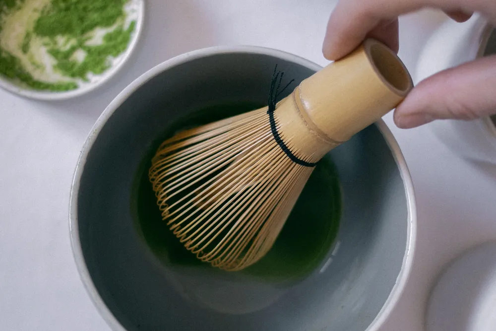 A bamboo whisk being used to prepare bright green matcha in a dark ceramic bowl.