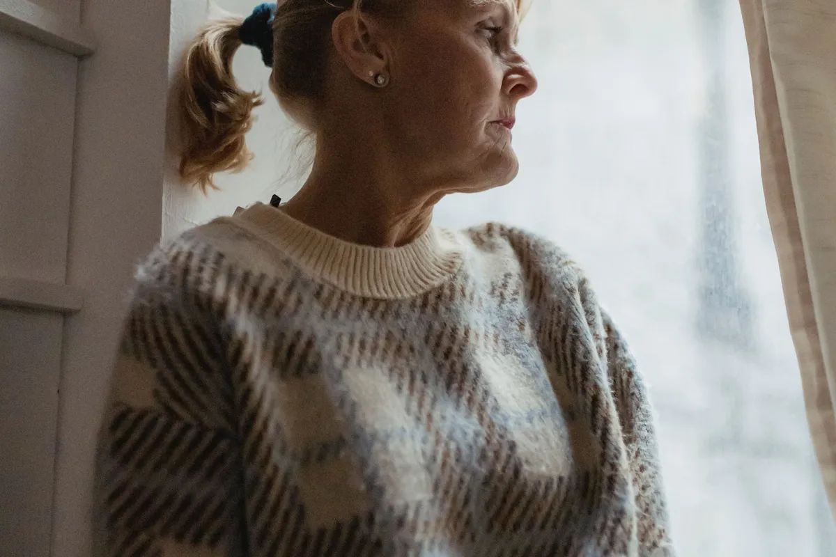 A woman in her fifties seen in profile beside a bright window, her jaw and neck in soft light.