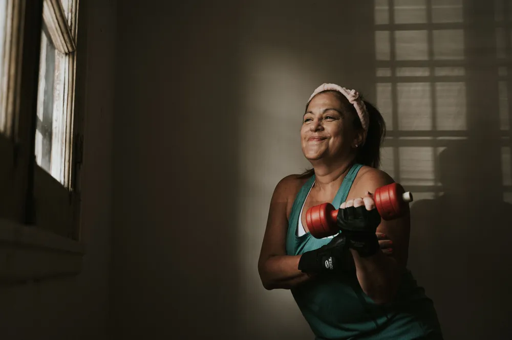 A middle-aged woman in a teal top smiling while lifting a red dumbbell indoors.