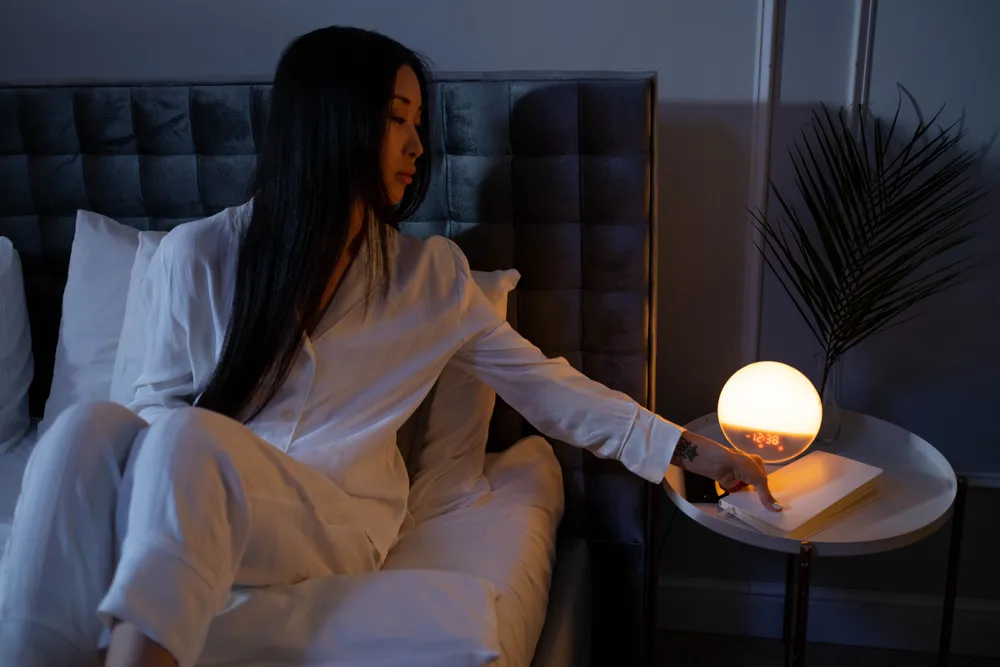 A woman in white pajamas reaching for a small glowing lamp on a nightstand in a dark bedroom.