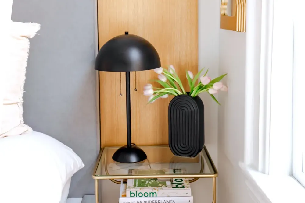 A tidy bedside table with a lamp, a small vase of flowers and a couple of books by a bright window.