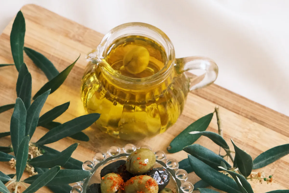 A glass jug of olive oil and a dish of green olives on a wooden board with fresh olive branches.