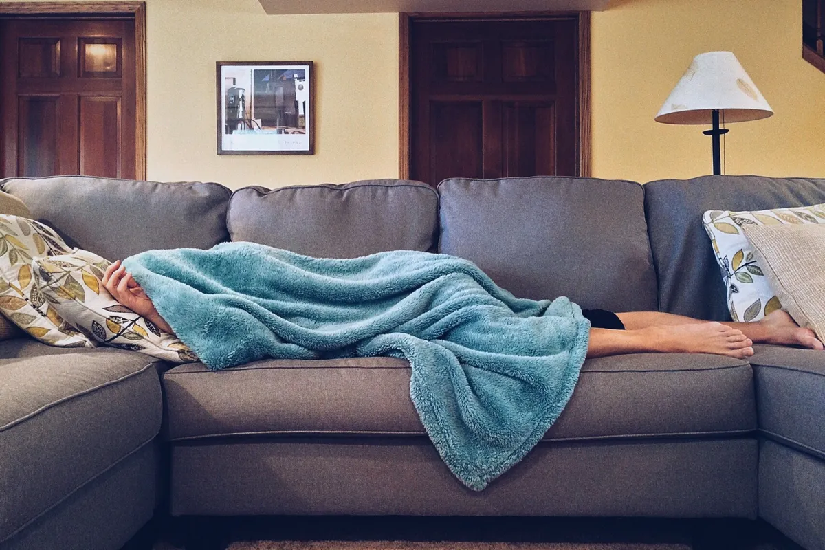 A person napping on a grey sofa under a blue blanket in a daylit living room.