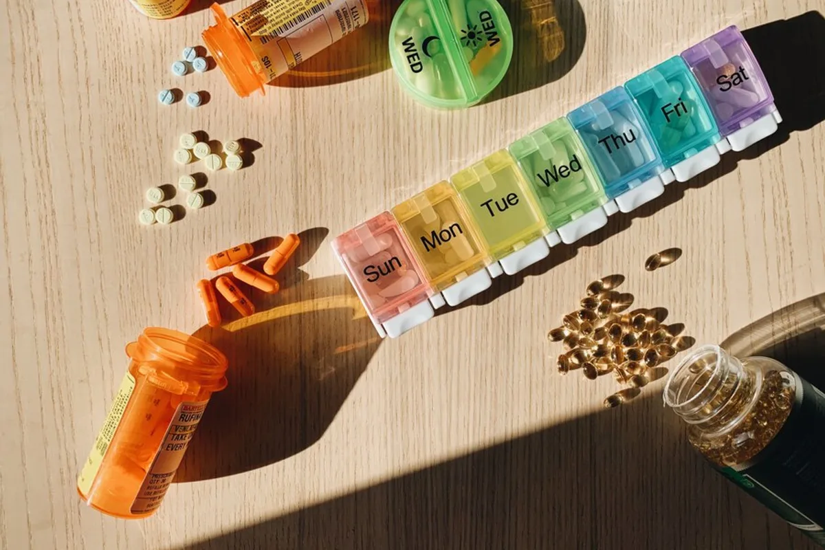 A weekly pill organiser on a wooden table beside orange prescription bottles and spilled supplement capsules.