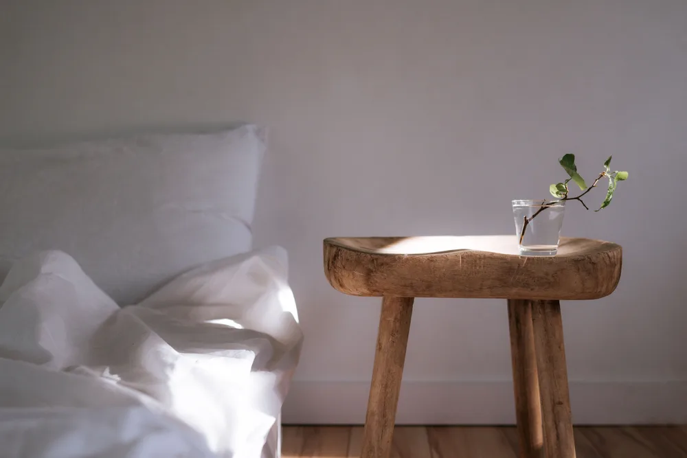 White pillows on a bed beside a small wooden bedside table with a sprig of greenery in soft daylight.