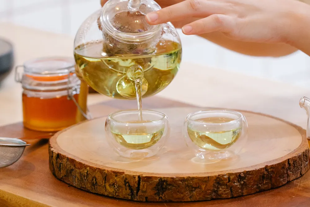 Hands pouring golden herbal tea from a glass teapot into two small glass cups beside a jar of honey.