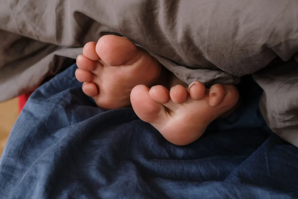 Two bare feet poking out from under a dark blanket in a dim bedroom at night.