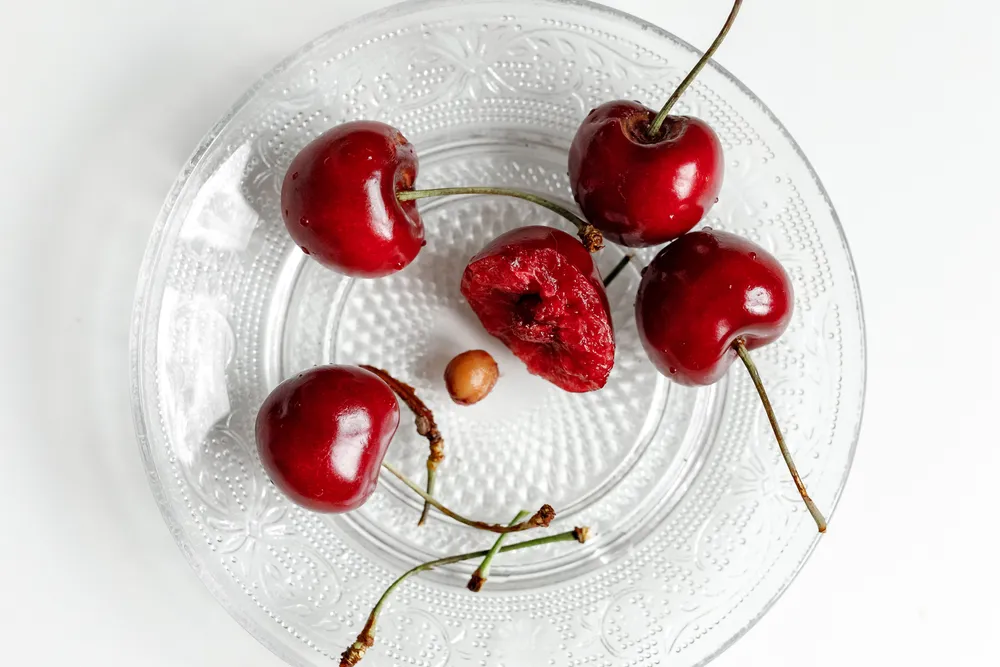 Fresh dark red cherries and one dried cherry arranged on a pressed glass plate.