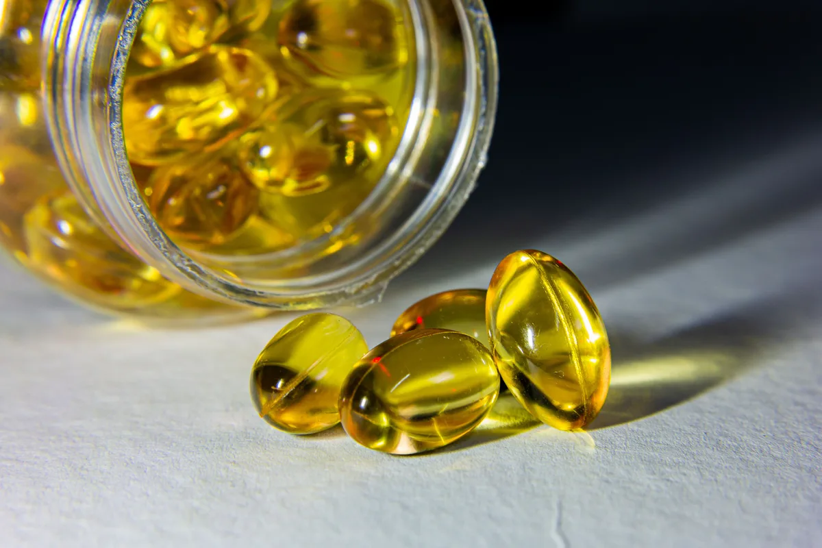 Golden vitamin D softgel capsules spilling from a tipped-over glass jar onto a pale surface.