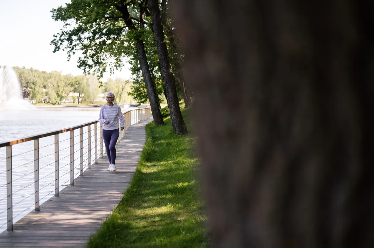 A woman in her fifties walking along a sunny lakeside boardwalk lined with trees.
