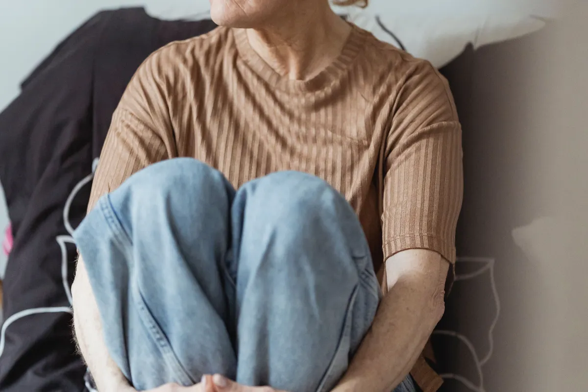 A woman sitting with her arms wrapped around her bent knees on a sofa, seen from the chin down.