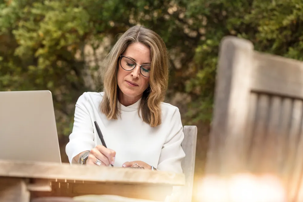 A woman in her forties wearing glasses, writing on a notepad beside an open laptop in daylight.