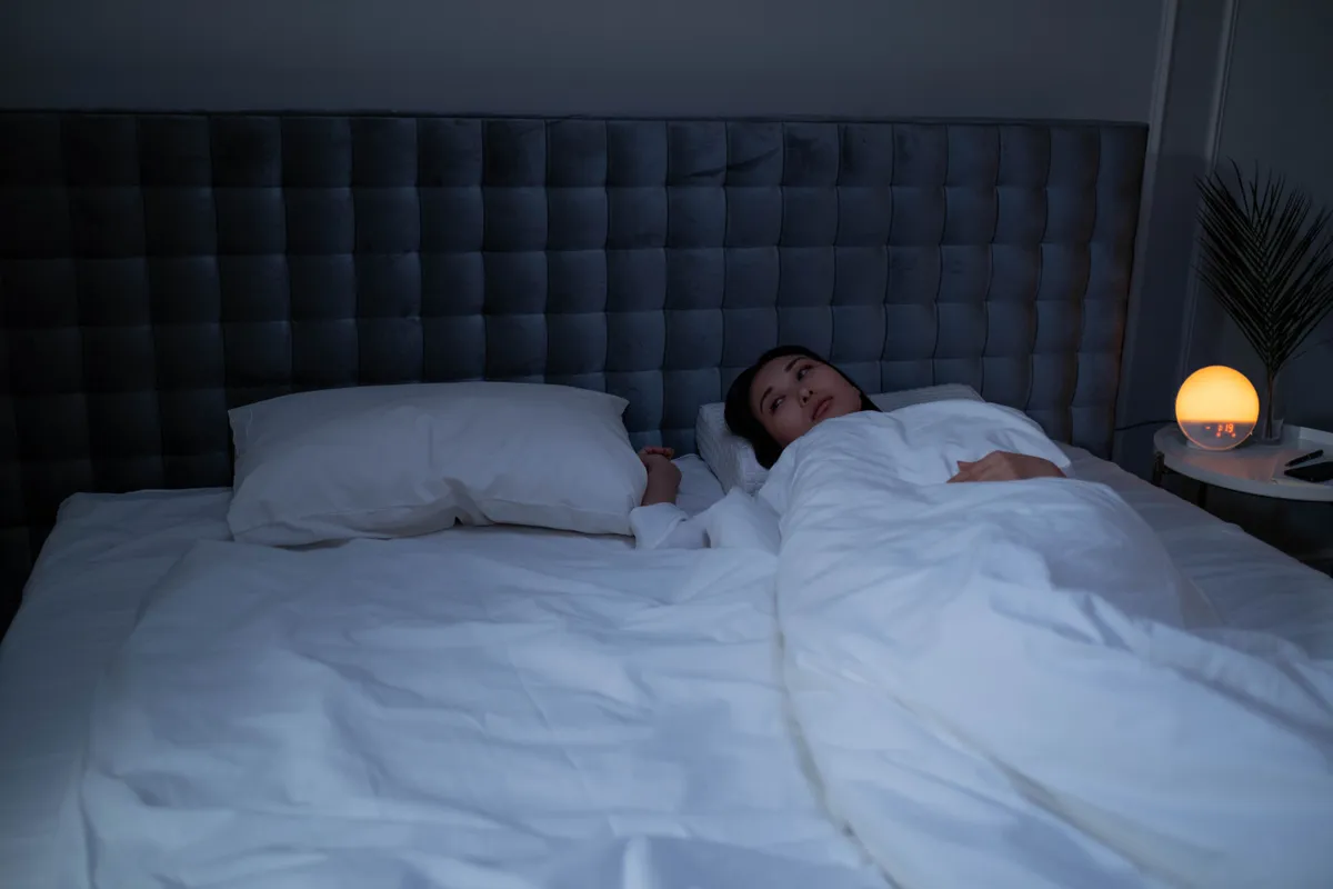 A woman lying awake in bed in a dark bedroom, lit only by a small warm bedside lamp.
