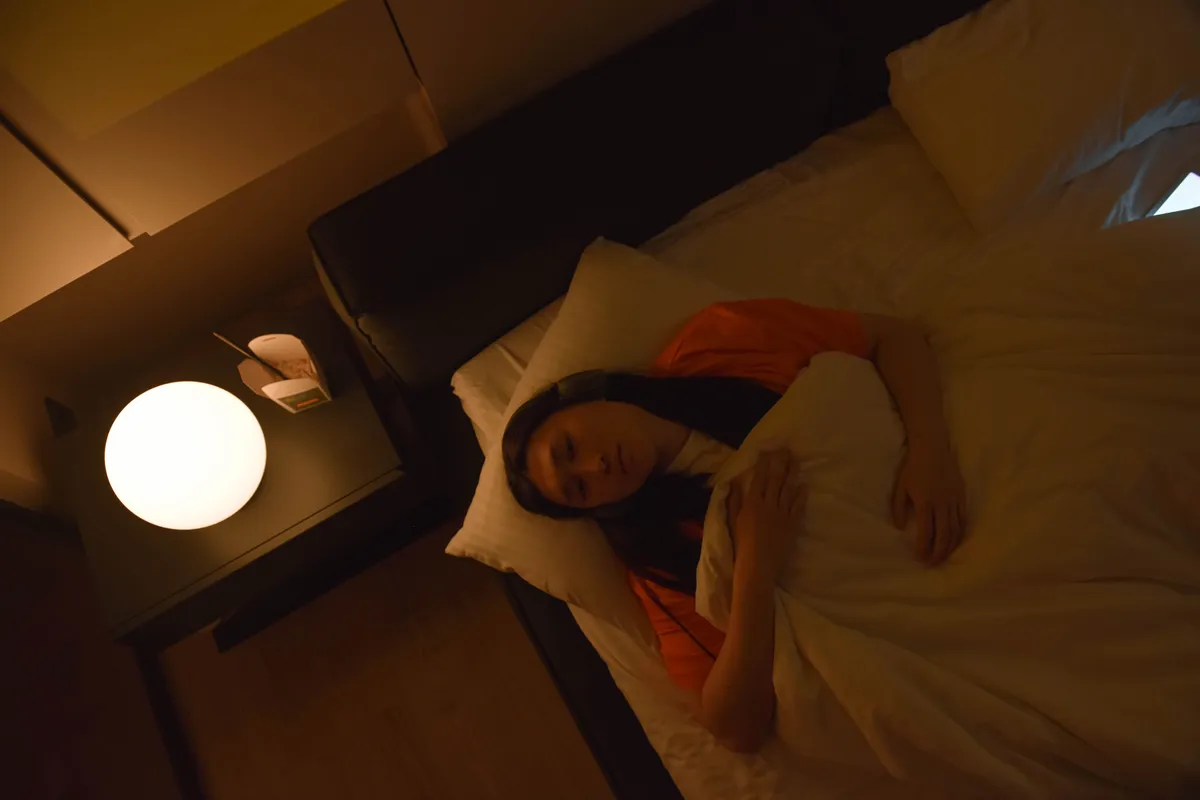A woman lying awake in a dark bedroom, lit by a single round bedside lamp.