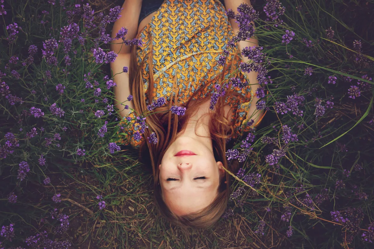 A woman lying peacefully in a lavender field, eyes closed, arms relaxed at her sides.