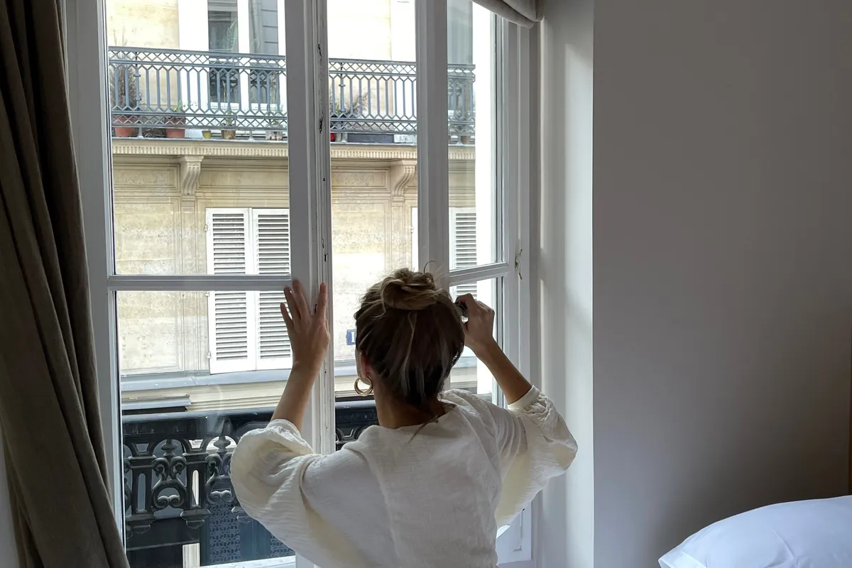 A woman seen from behind opening a tall bedroom window as daylight comes in.