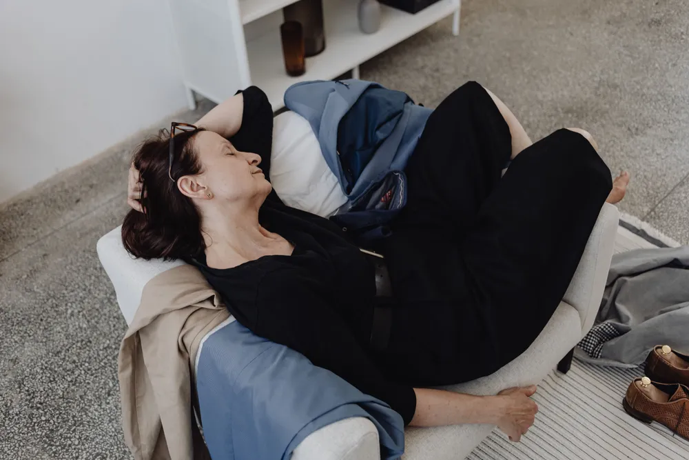 A woman lying down indoors with her eyes closed, still fully dressed, resting on a cushion.