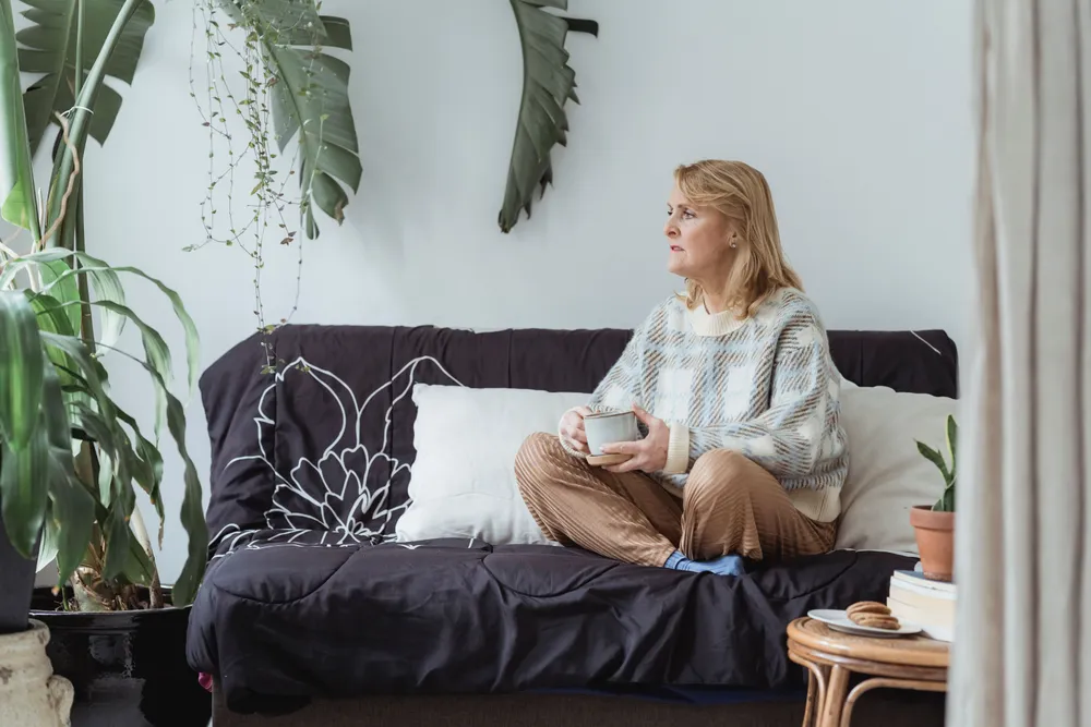 A woman sitting on a sofa in a plant-filled living room, holding a warm drink in both hands.