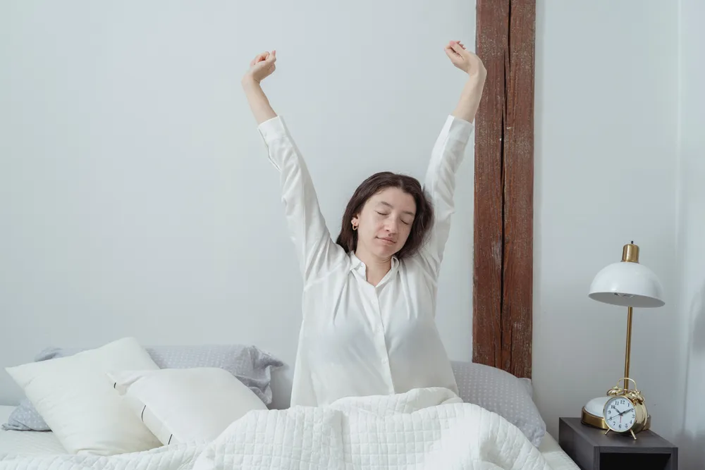 A woman stretching her arms above her head in bed, in a bright bedroom in the morning.