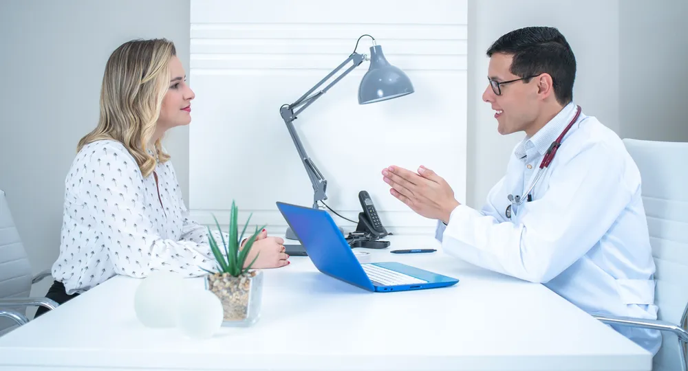 A woman in her fifties talking with a doctor across a desk in a bright consulting room.
