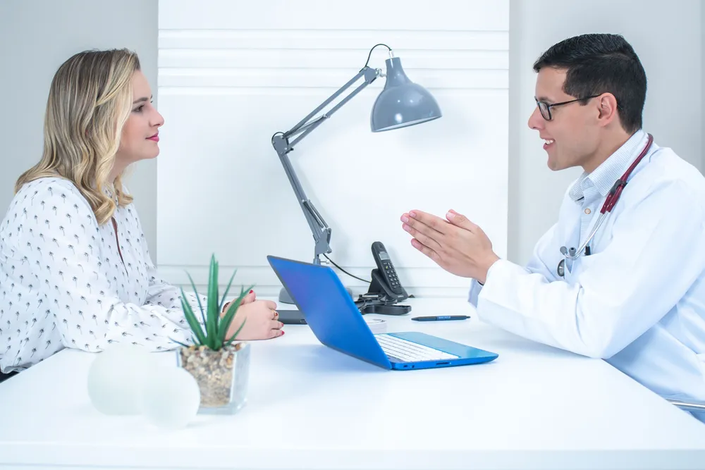 A woman sitting across a desk from a doctor in a white coat during a consultation.