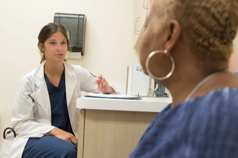 A woman with short hair and hoop earrings, seen from behind, talking with a female doctor in a clinic room.