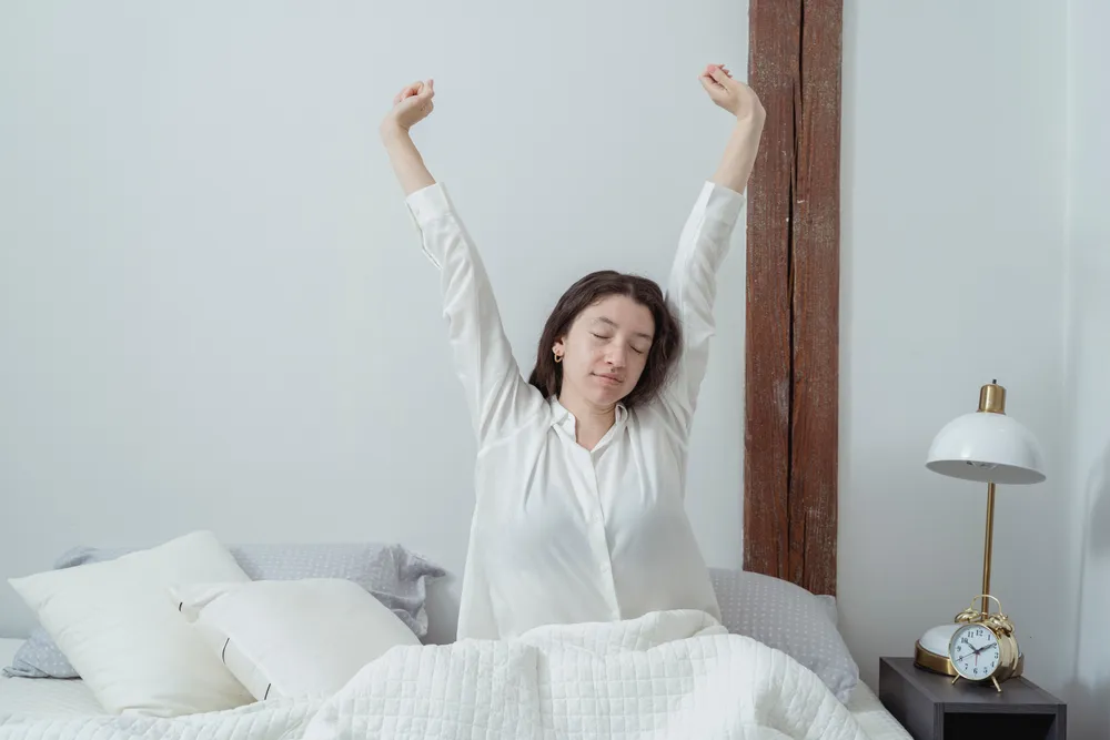 A woman sitting up in bed stretching both arms overhead in a bright white bedroom, an alarm clock on the bedside table.